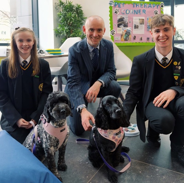 A photo of brother and sister pupils Paul and Margot Kennedy and their headteacher Huw Brophy from St Thomas More RC College in Denton visiting the proton beam therapy centre at The Christie to meet resident therapy dogs Lilo and Luna.