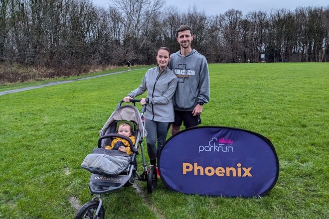 A photo of Christie patient Zac Hinchcliffe and his wife Danni in running clothes, with their baby son in a push chair. The 3 are in a park at a parkrun event, a sign reads 'parkrun Phoenix'.