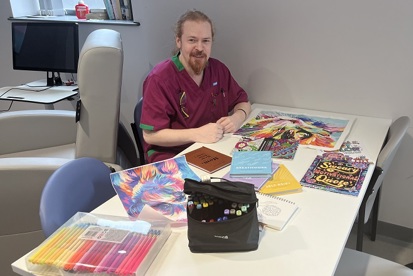 A photo of Ivan Cobham, activity coordinator at The Christie, sitting at a table with colouring pens and art supplies.