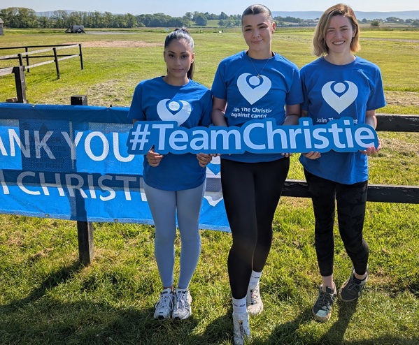 A photo of Christie nurses Rasleen Sharma, Rhiana Smith and Abbie Gunton wearing Christie Charity t-shirts and holding a sign that reads '#TeamChristie'; they are standing in front of a field.