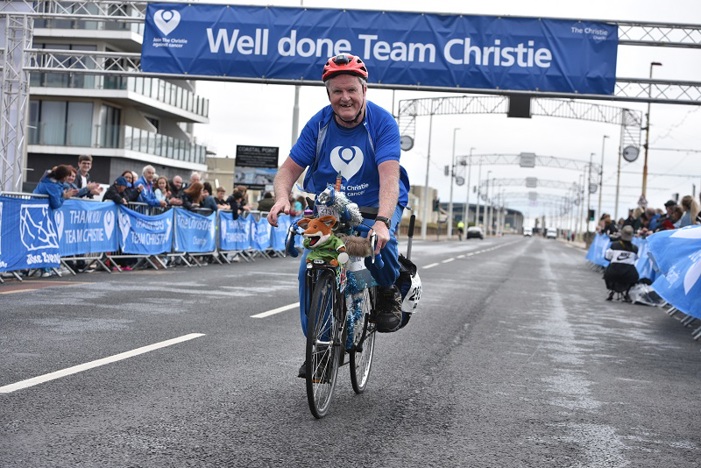A photo of Tony O'Dwyer riding a bike along the Blackpool promenade with a Basil Brush toy on the front of his bike; a sign above him reads "Well done Team Christie".