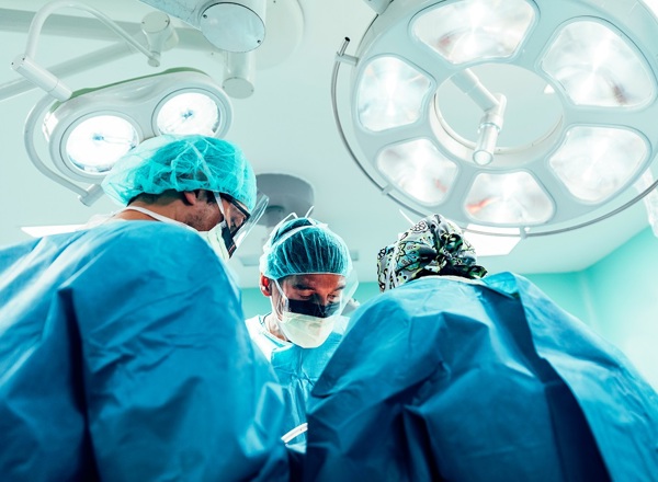 A photo of 3 doctors in scrubs, hairnets and facemasks standing underneath some surgery lights.