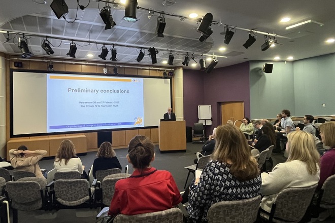 A photo of Simon Osbert from the OECI presenting to a group of people in The Christie auditorium; the screen read 'Preliminary conclusions: Peer review 26 and 27 February 2025, The Christie NHS Foundation Trust'.