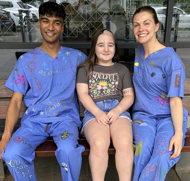 A photo of Christie radiographers Kiyan and Dayna wearing their decorated scrubs and sitting on a bench with young Christie patient Esmai Evans.