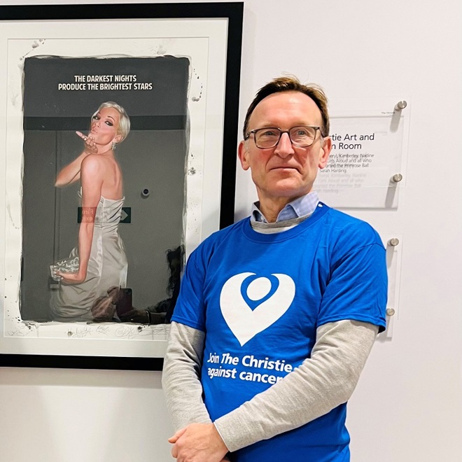 A photo of Dr Sacha Howell standing outside The Christie art room in front of photo of Sarah Harding which reads 'The darkest nights produce the brightest stars'.