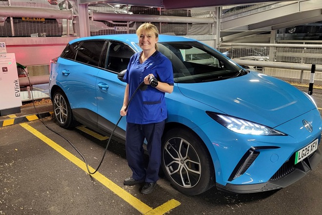 A photo of Christie at Home nurse Sarah Richardson standing next to a blue electric MG4 car.