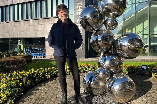 A photo of Christie patient Adele Adams standing in front of the sculpture outside of The Christie entrance.