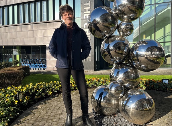 A photo of Christie patient Adele Adams standing in front of the sculpture outside of The Christie entrance.