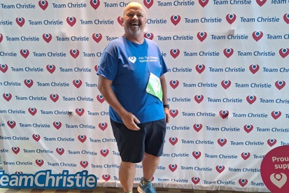 A photo of Mark Love, Christie fundraiser, wearing a Christie Charity t-shirt and standing in front of a Team Christie background.
