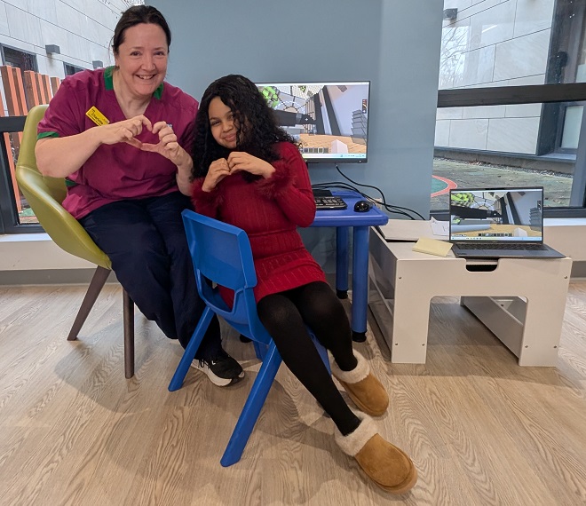 A photo of Christie patient Rama with Health Play Specialist Charlotte Cooper sitting in front of a computer screen with the proton beam therapy centre at The Christie built in Minecraft.