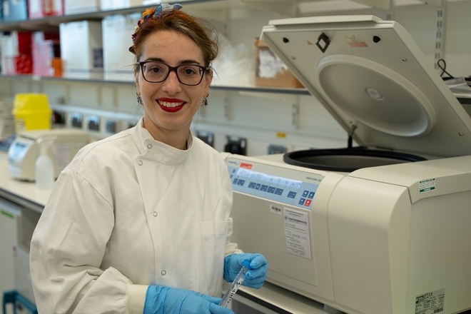 A photo of Dr Sara Valpione, a consultant at The Christie, wearing a white coat and rubber gloves and standing in front of a centrifuge.