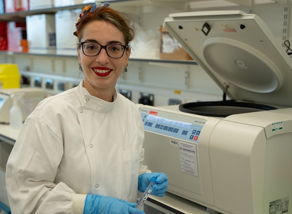 A photo of Dr Sara Valpione, a consultant at The Christie, wearing a white coat and rubber gloves and standing in front of a centrifuge.