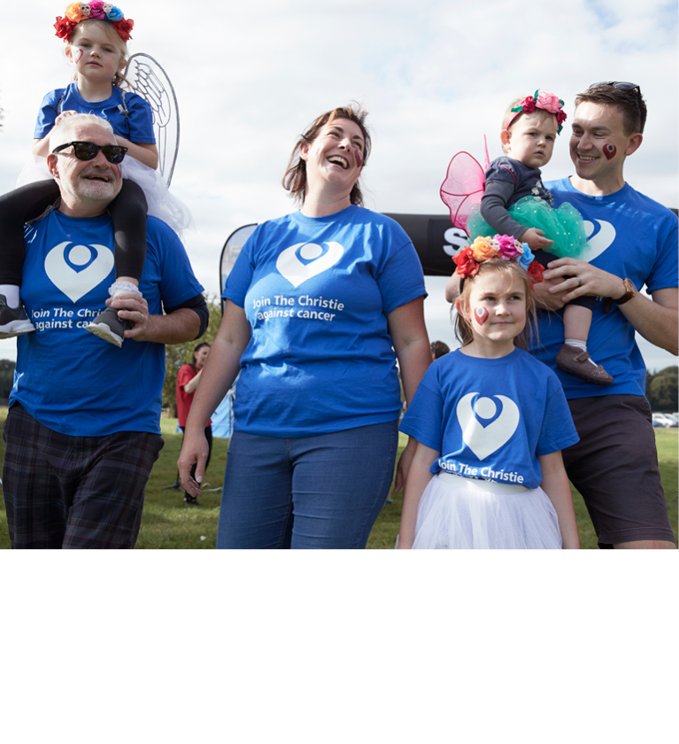A photo of a family of 3 adults and 3 children. An older man on the left has a young girl with a flower crown riding on his shoulders; a woman in the middle is laughing and has her hand on a girl's arm, and a man on the right is carrying a toddler. Everyone is wearing The Christie Charity t-shirts.
