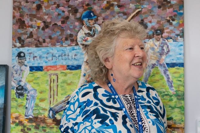 A photo of Pat Mountford, the artist in residence at The Christie, standing in front of a painting by a Christie patient.