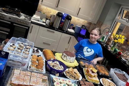 A photo of Lily Huggins standing in a kitchen and wearing a Christie t-shirt behind a selection of homebaked cakes.