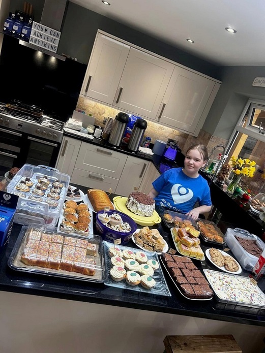 A photo of Lily Huggins standing in a kitchen and wearing a Christie t-shirt behind a selection of homebaked cakes.