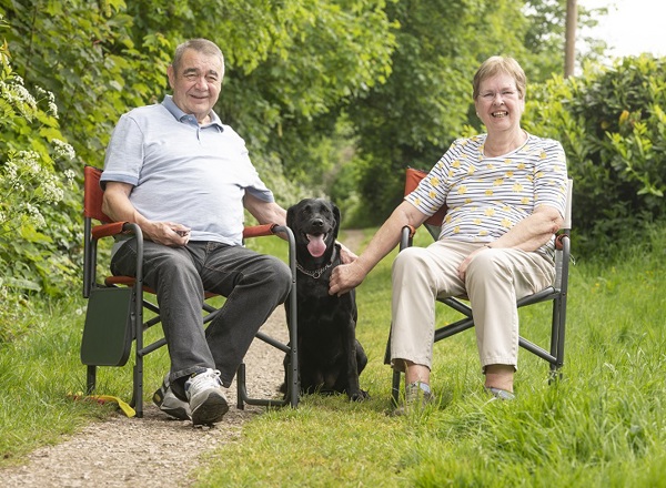 A photo of Christie patient Eileen Unsworth sitting in a chair on the right with her husband Peter on the left and their black Labrador Annie sitting between them.