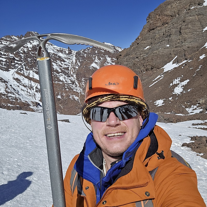 A photo of Christie fundraiser Matt Heywood on top of snowy Mount Toubkal wearing a hard hat and holding an ice pick.