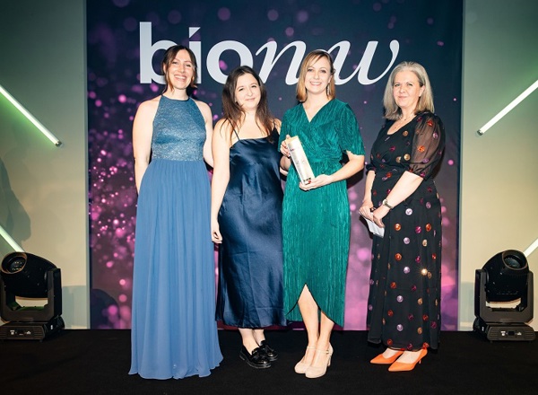 A photo of 4 female members of the Experimental Cancer Medicine Team at The Christie accepting their Bionow 2023 Social Impact Award at the Bionow Awards ceremony, with a purple background reading Bionow.