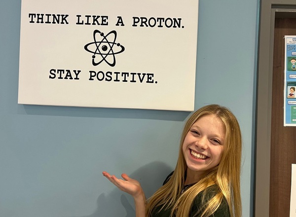 A photo of Eve Wilson, a former patient at the proton beam therapy centre at The Christie, stands in front of a sign that reads 'Think like a proton. Stay positive."